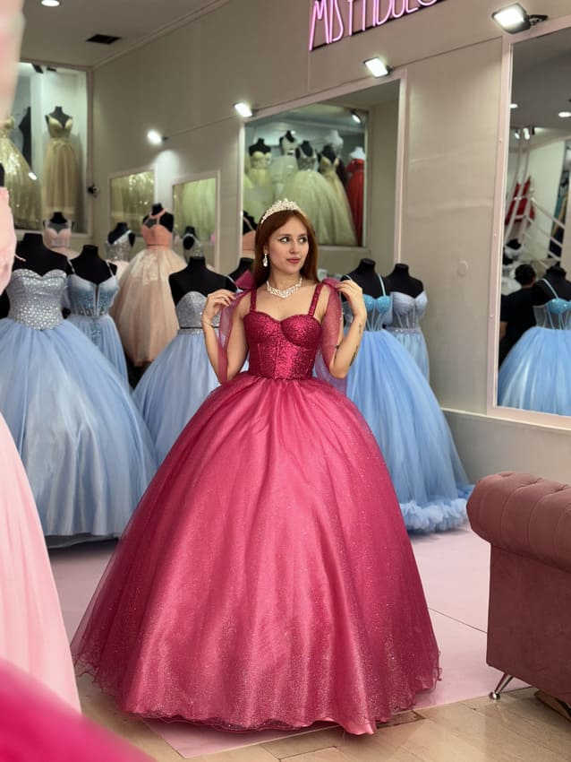 Vestido de 15 fucsia estilo princesa con corset brillante y breteles, falda amplia de tul con volumen, exhibido en tienda de vestidos de fiesta.