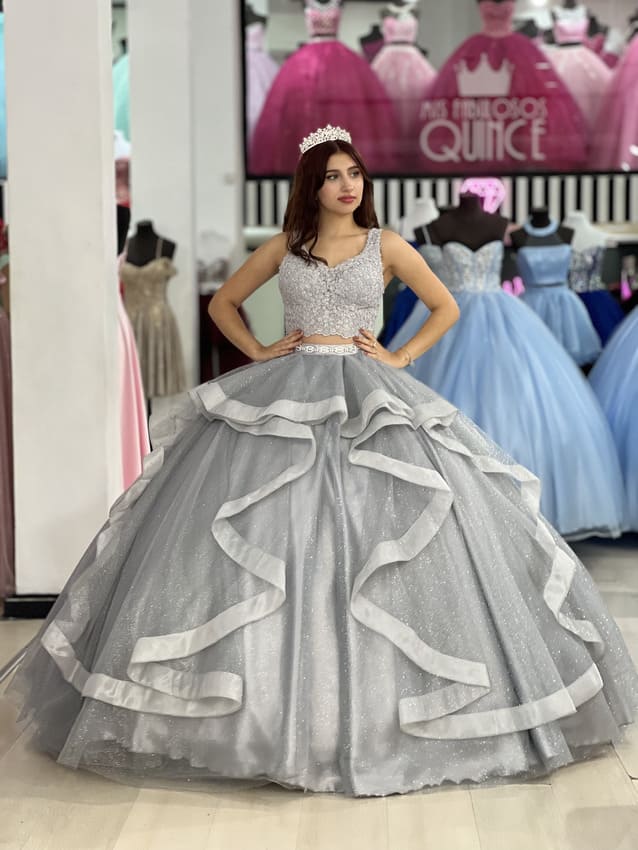 Vestido de 15 gris plateado con falda amplia de volados y corset bordado, estilo princesa, exhibido en una tienda de vestidos de fiesta.