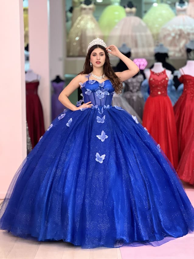 Vestido de quince azul con falda voluminosa de tul brillante y aplicaciones de mariposas, usado por una joven con tiara en una tienda de vestidos.