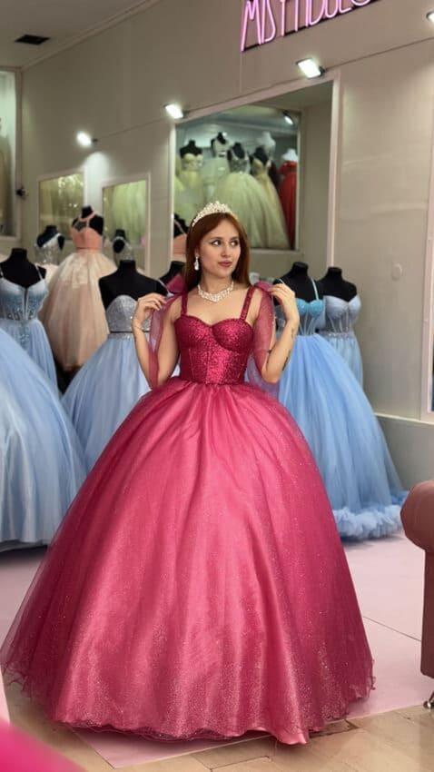 Vestido de 15 fucsia estilo princesa con corset brillante y breteles, falda amplia de tul con volumen, exhibido en tienda de vestidos de fiesta.