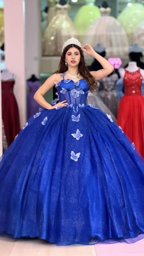 Vestido de quince azul con falda voluminosa de tul brillante y aplicaciones de mariposas, usado por una joven con tiara en una tienda de vestidos.